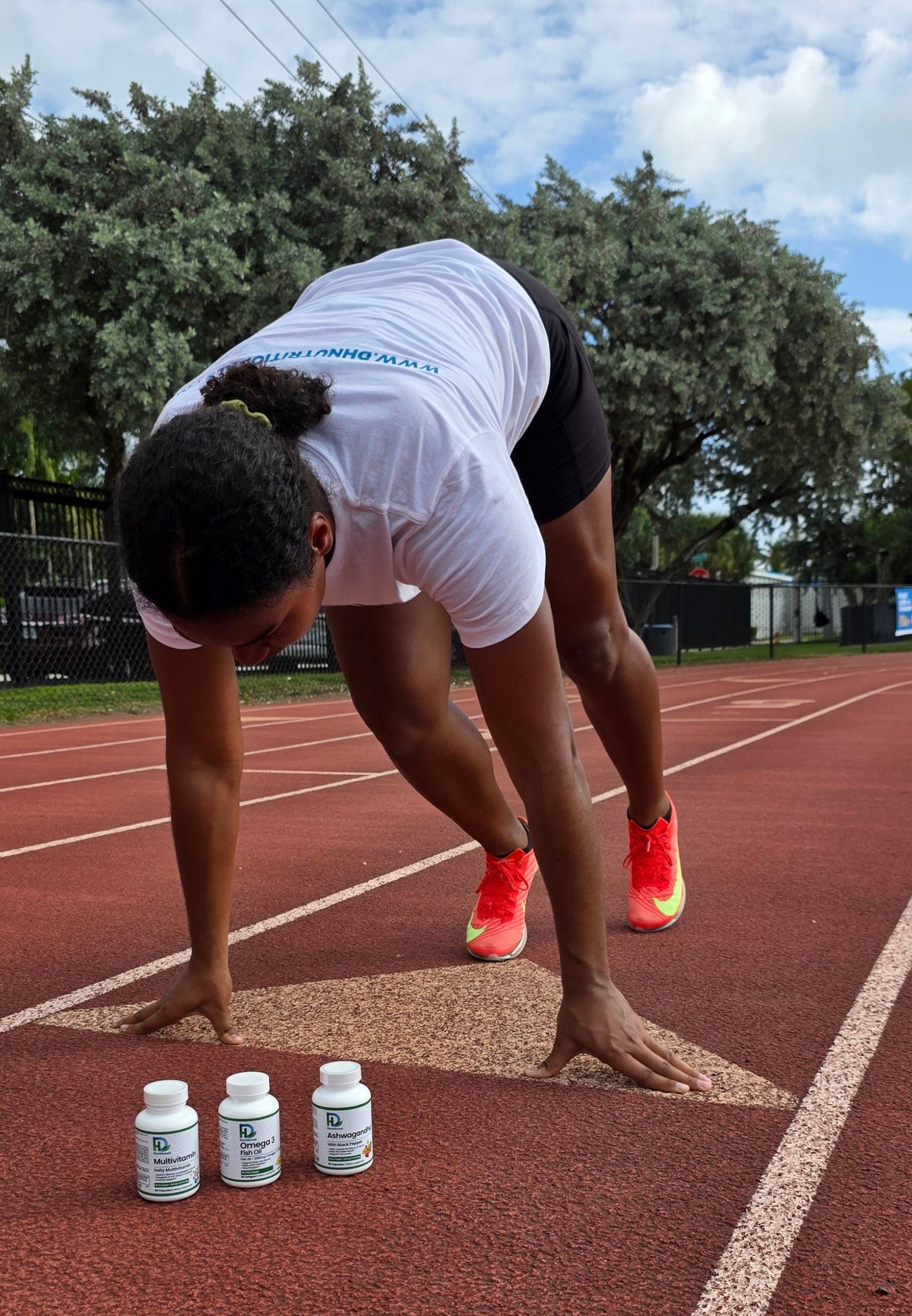 A person in a white t-shirt and black shorts crouches on a red running track, with three white supplement bottles placed on the track in the foreground. Trees and a blue sky are visible in the background.